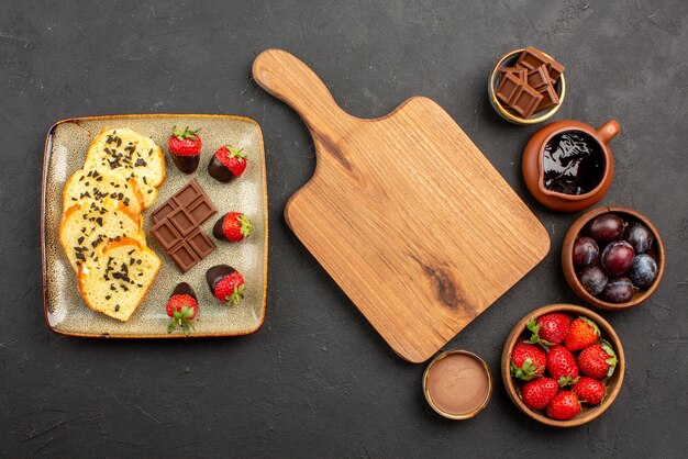 Top view cake and strawberries wooden board between pieces of cake with chocolate on the left and bowls with strawberries berries and chocolate sauce on the right side of the table