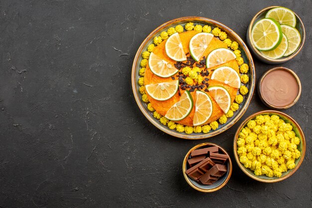 Top view cake with citrus fruits appetizing cake with chocolate and citrus fruits next to the bowls of slices of lime chocolate and chocolate cream on the black table