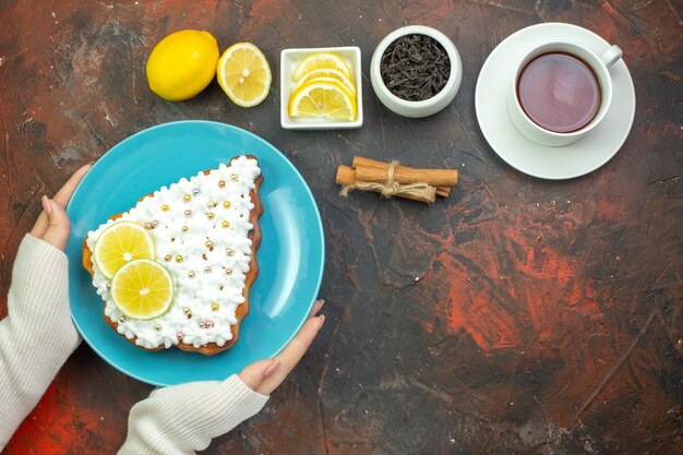 Top view cake with lemon on blue plate in female hand lemon slices in bowl cup of tea cinnamon sticks on dark red background