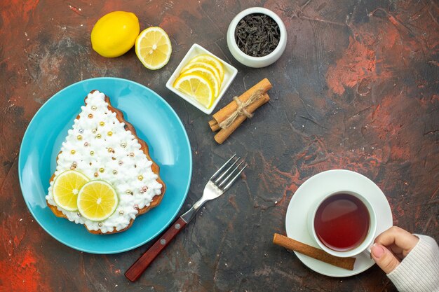 Вид сверху торт с лимоном на синей тарелке, ломтики лимона в миске, вилка, чашка чая в женской руке, палочки корицы на темно-красном фоне
