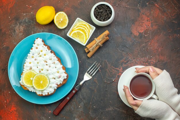 Top view cake with lemon on blue plate lemon slices in bowl fork cup of tea in woman hands cinnamon sticks on dark red background