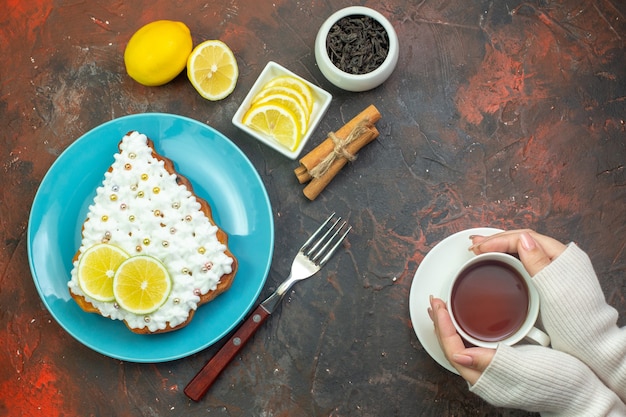 Top view cake with lemon on blue plate lemon slices in bowl fork cup of tea in woman hands cinnamon sticks on dark red background