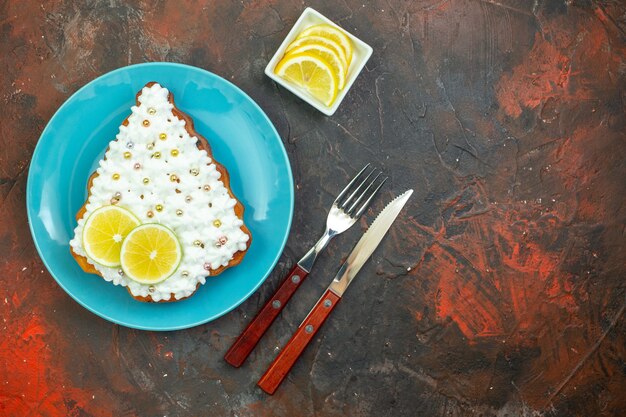 Вид сверху торт с лимоном на синей тарелке, ломтики лимона в миске, нож и вилка на темно-красном фоне