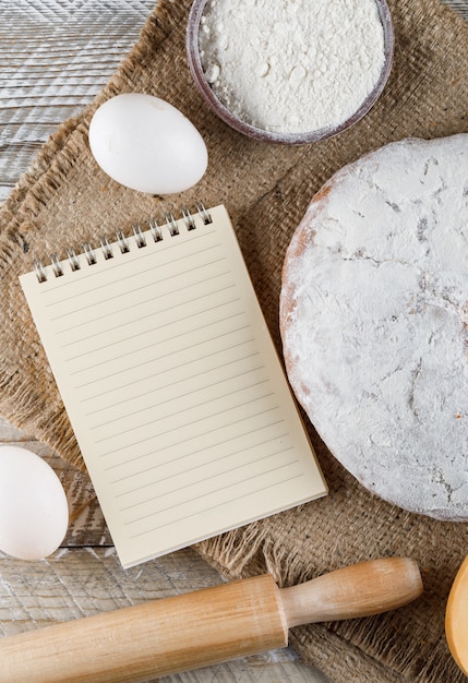 Top view cake with notepad, eggs, rolling pin on sack cloth and wooden surface. vertical