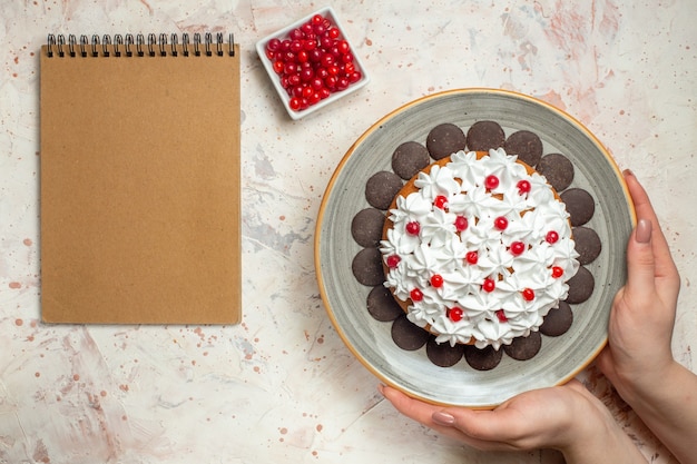 Top view cake with pastry cream and chocolate in female hand berry in bowl and empty notebook