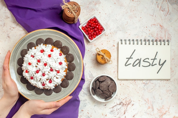Торт вид сверху с кремом для выпечки на серой овальной тарелке в руке женщины. вкусное слово, написанное в блокноте на белом столе
