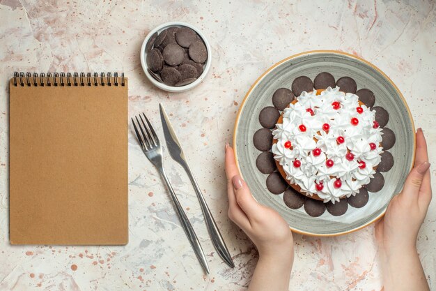 ボウルフォークとディナーナイフノートブックの女性の手のチョコレートのプレートにペストリークリームとトップビューケーキ