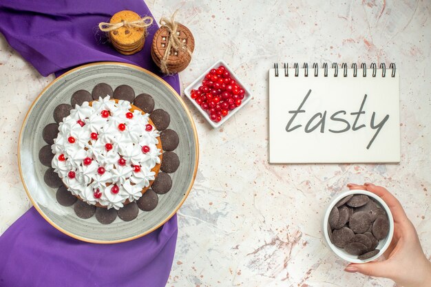 top view cake with pastry cream on plate purple shawl cookies tied with rope berries in bowl chocolate bowl in female hand tasty written on notepad on white table