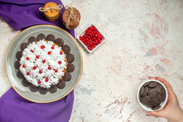 プレート上のペストリークリームとトップビューケーキ白いテーブルの上の女性の手でボウルチョコレートボウルのロープベリーと結ばれた紫色のショールクッキー