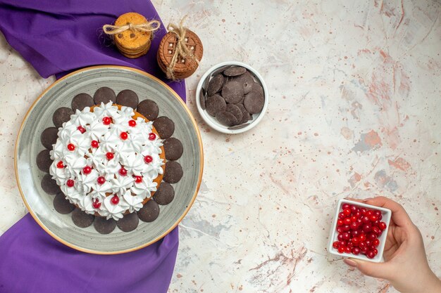 Вид сверху торт с кондитерским кремом на тарелке фиолетовое печенье с шалью, перевязанное веревкой, шоколад в миске ягодная миска в женской руке на белом столе