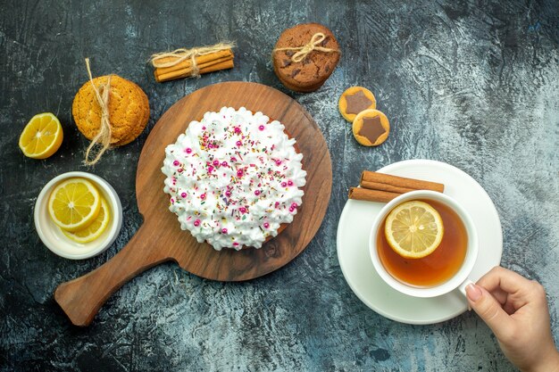 木のサービングボードクッキーシナモンスティックグレーのテーブルの上の女性の手にお茶のカップとペストリークリームとトップビューケーキ