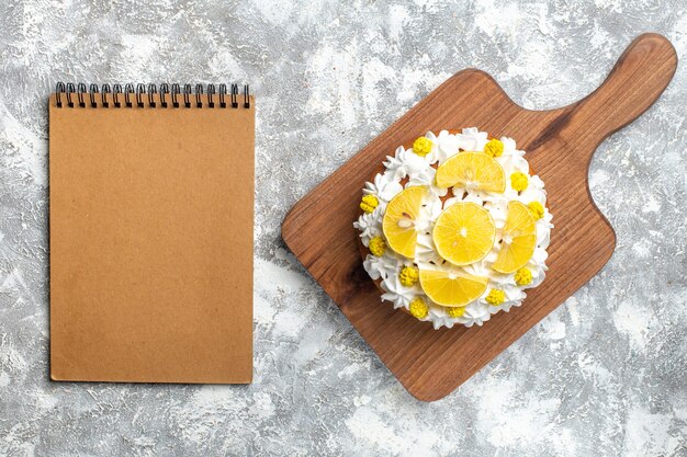 Top view cake with white cream and lemon slices on cutting board and empty notebook