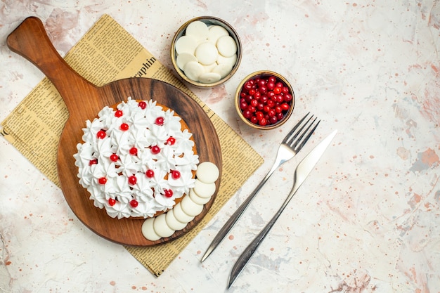 Top view cake with white pastry cream on cutting board on newspaper fork and dinner knife bowls with white chocolate and berries on light grey table