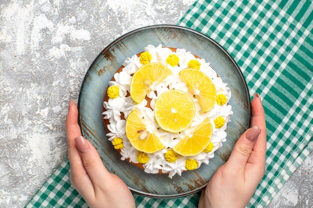 Вид сверху торт с белым кондитерским кремом и ломтиками лимона на круглой тарелке в руке женщины