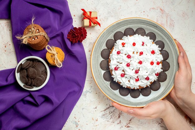 Top view cake with white pastry cream on plate in woman hand cookies tied with rope chocolate bowl on purple shaw