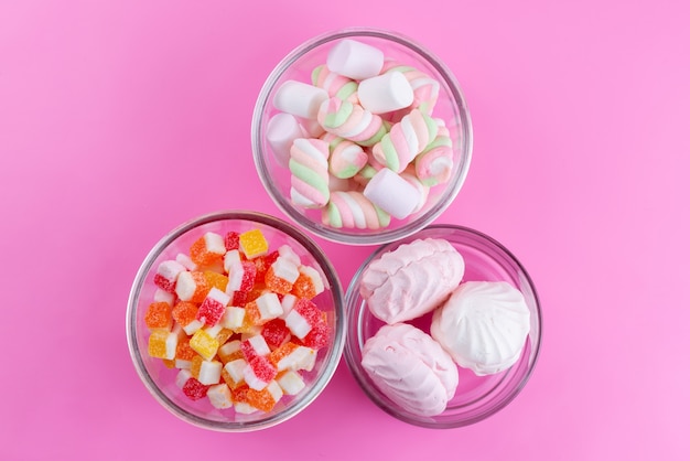 A top view candies and meringues along with marshmallows inside glass bowls on pink