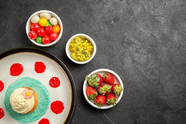 Top view candies on the table plate of cake with colorful sauces next to the bowls of strawberries herbs and candies on the table