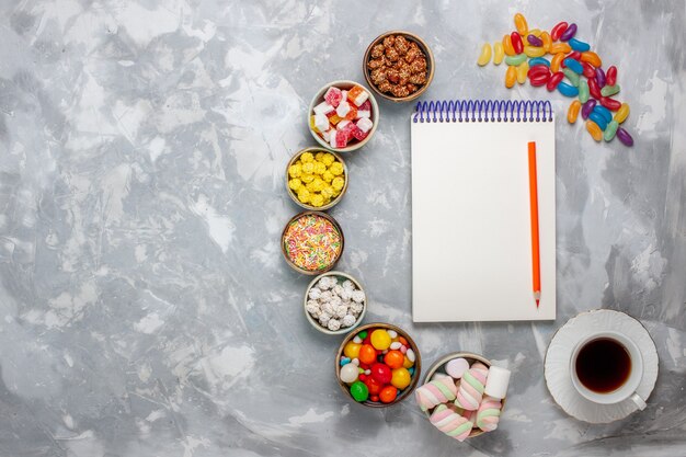 Вид сверху конфетной композиции разноцветные конфеты с зефирным блокнотом и чаем на белом столе сахарная конфета конфитюр сладкий конфитюр