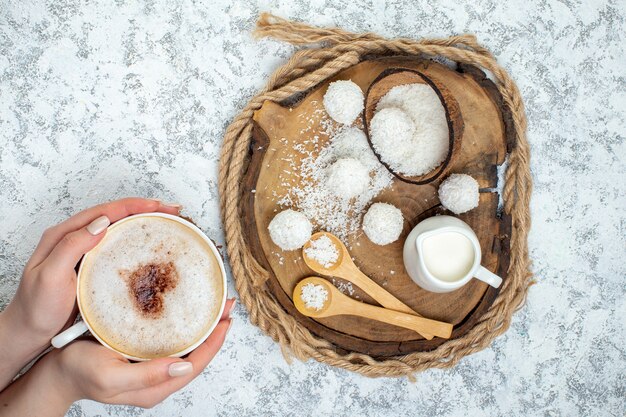 Top view cappuccino cup in female hand coconut balls milk bowl spoons on wood board on grey surface