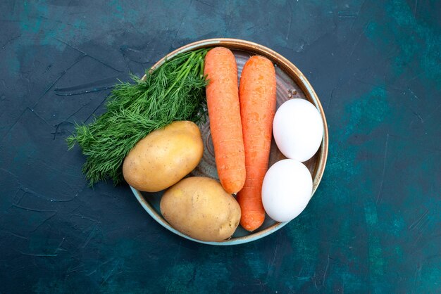 Top view carrots and potatoes with eggs and greens on dark-blue desk.