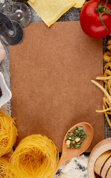 Top view of carton sheet and italian raw pasta of different types and shapes with fresh tomatoes and cutter roller knife on black background