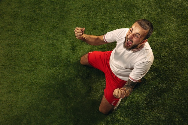 Top view of caucasian football or soccer player on green background of grass. young male sportive model training, practicing. kicking ball, attacking, catching. concept of sport, competition, winning.