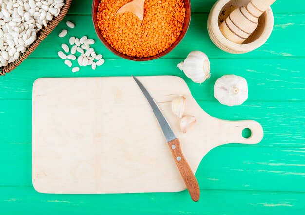 Top view of cereals and legumes in bowls and a wooden cutting board with a knife on green background