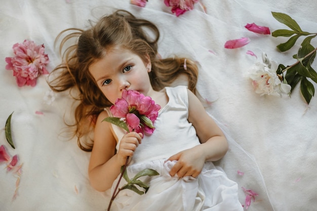 Vista dall'alto dell'affascinante ragazza piccola che tiene in mano una peonia rosa tenue sdraiata su un lenzuolo bianco, circondata da fiori freschi, seriamente guardando dritto