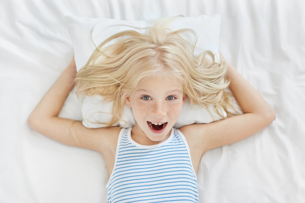 免费照片顶视图的欢快的小女孩的金发,feckles蓝眼睛穿脱下睡衣躺在白色的枕头和床单在床上,玩和笑,不想白天的小睡