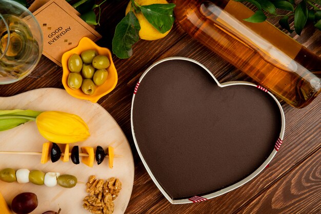 Top view of cheese set as cheddar and parmesan with olive walnut grape and flower on cutting board with heart-shaped box white wine lemon and good everyday card on wooden background