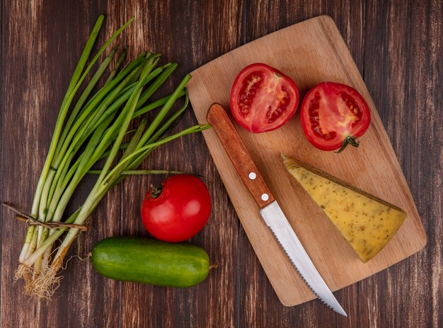 Top view cheese slices with tomatoes and knife on a stand with cucumber and green onions on wooden background