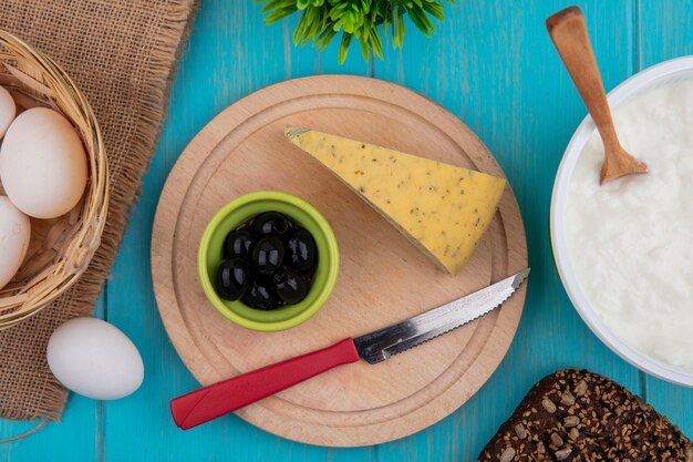 Вид сверху сыр с оливками и ножом на подставке с йогуртом в миске на бирюзовом фоне