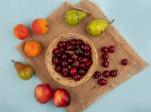 Top view of cherries in basket and pattern of pears apricots peaches cherries on sackcloth on blue background