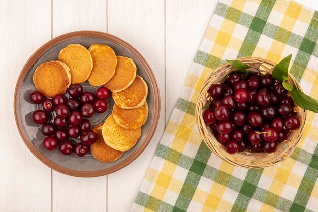 Top view of cherries in basket on plaid cloth and plate of pancakes and cherries on wooden background