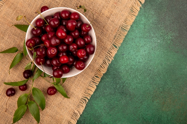 Top view of cherries in bowl with leaves on sackcloth on green background with copy space