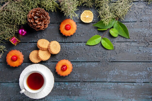 Top view cherry cupcakes fir-tree branches slice of lemon a cup of tea biscuits and leaves on dark wooden table with copy space