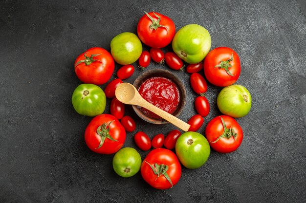 Top view cherry red and green tomatoes around a bowl with ketchup and a wooden spoon on dark ground with copy space