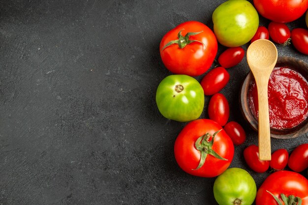 Top view cherry red and green tomatoes around a bowl with ketchup and a wooden spoon on dark table with copy space