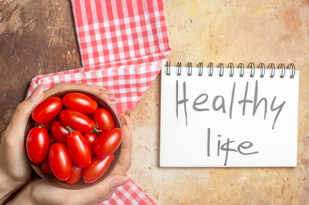 Top view cherry tomatoes in wooden bowl in female hands a kitchen towel healthy life written on notepad on amber background