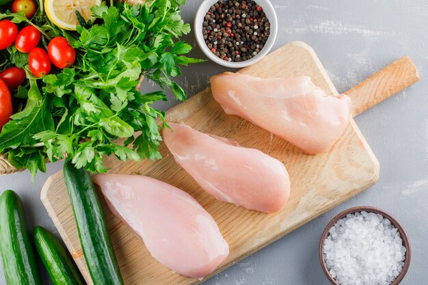 Top view chicken breast on a cutting board with cucumber, greens, salt, pepper on gray surface