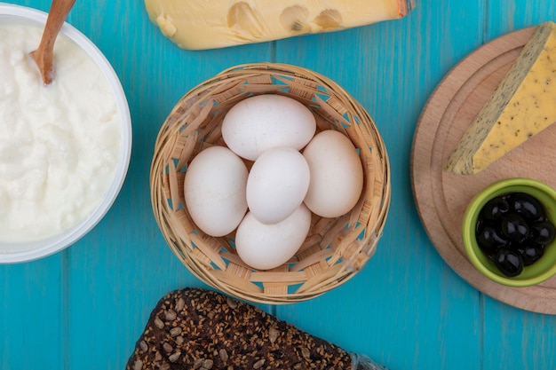 Top view chicken eggs in a basket of cheese with black bread and yogurt in a bowl on a turquoise background