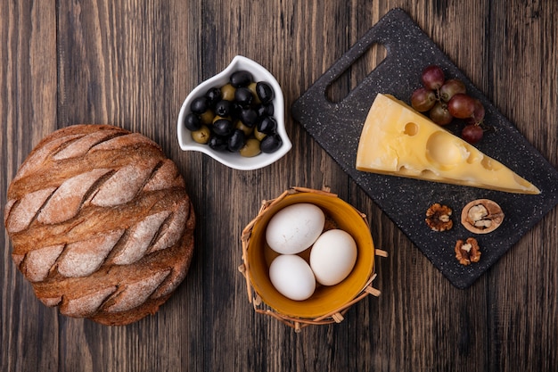 Top view chicken eggs in a basket of olives in a saucer with maasdam cheese on a stand with black bread on a wooden background