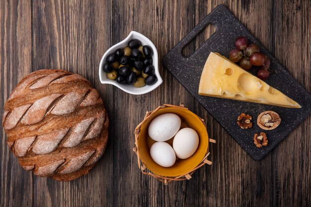 Top view chicken eggs in a basket of olives in a saucer with maasdam cheese on a stand with black bread on a wooden background