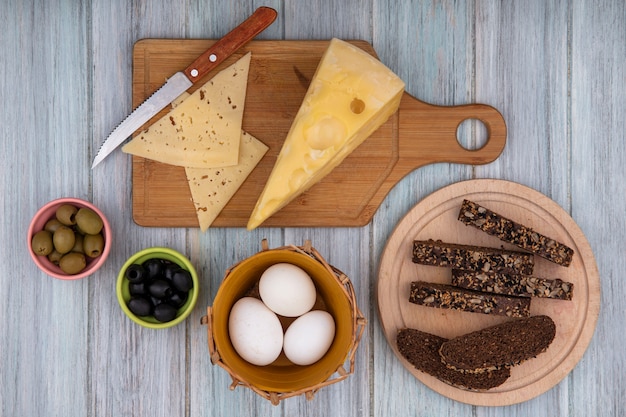 Free photo top view chicken eggs in a basket with slices of black bread on a stand with cheeses on a cutting board and a knife  on a gray background