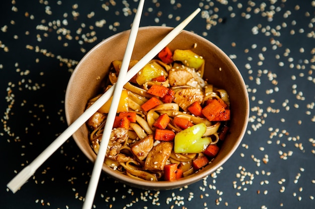 Top view chicken noodles with vegetables in plate with chopsticks and sesame seeds on black background