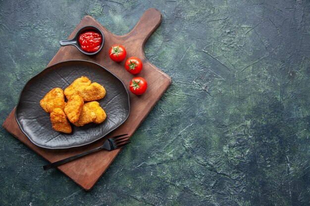 Top view of chicken nuggets on a black plate and fork on wooden board tomatoes ketchup on the right side on dark surface