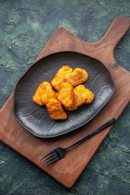 Top view of chicken nuggets on a black plate and fork on wooden cutting board on dark surface