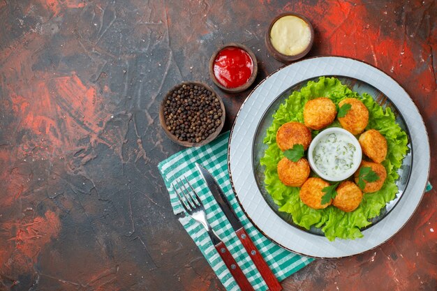 Top view chicken nuggets lettuce and sauce on plate sauces and black pepper in small bowls knife and fork on dark table free space