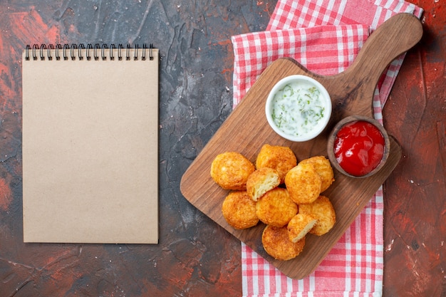 Top view chicken nuggets on wood board with sauces red white
checkered kitchen towel notebook on dark table