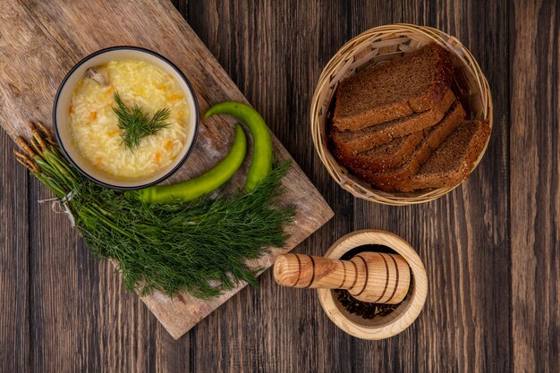 Top view of chicken orzo soup in bowl and peppers with dill on cutting board and basket of rye bread slices with black pepper on wooden background